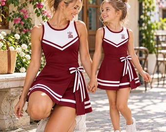 May include: Two matching sleeveless athletic dresses in maroon and white. Each dress features a white bodice, a maroon skirt with white horizontal stripes, and a side-tie bow detail. A small logo is printed on the chest of each garment.