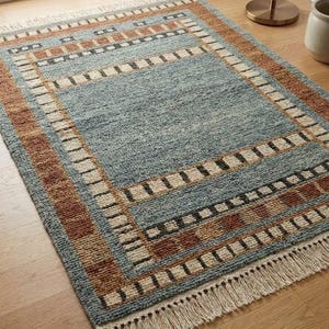 May include: A rectangular area rug featuring a central blue field surrounded by a border of alternating brown, cream, and dark gray geometric blocks. The rug has a textured, woven surface and is finished with cream-colored fringe on both ends.