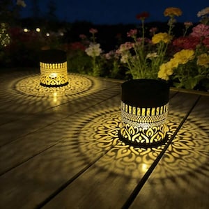 May include: Two cylindrical metal lanterns with a black exterior and intricate laser-cut patterns. The lanterns emit a warm yellow light, casting detailed, symmetrical floral-like shadow projections onto the wooden surface beneath them.