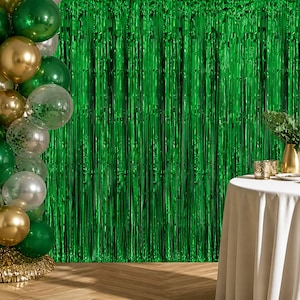May include: A metallic green fringe curtain backdrop paired with a cluster of green, gold, and clear confetti-filled balloons. A white tablecloth covers a round table holding two small gold glasses and a green plant in a gold vase.