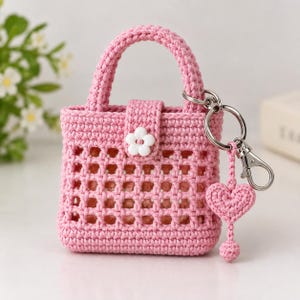 May include: A small pink crocheted handbag featuring a grid-like open weave pattern, a single top handle, and a central tab closure secured with a white flower-shaped button. A silver metal keychain with a small crocheted heart charm is attached.