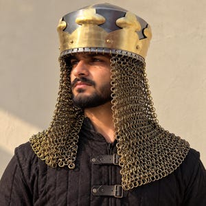 Könnte beinhalten: Ein Helm im mittelalterlichen Stil mit einer polierten Messingkrone mit gewelltem Rand und einer dunklen Metallkuppel. An der Basis ist eine lange, schützende Kettenhaube aus ineinandergreifenden Metallringen befestigt, die über die Schultern fällt.