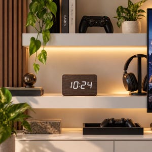 Puede incluir: Un reloj despertador digital rectangular con acabado de veta de madera oscura y una pantalla LED blanca brillante que muestra la hora 10:24. El reloj está colocado en un estante blanco, rodeado de varios objetos decorativos, plantas y aparatos.