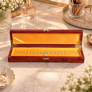 May include: A rectangular wooden jewelry box with a dark reddish-brown finish and a gold-colored metal latch. The interior is lined with bright yellow satin fabric and contains a delicate silver-toned chain bracelet with clear crystals.