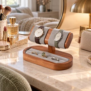 May include: A wooden watch and ring display stand featuring a horizontal bar with two grey velvet cushions for watches and a base with a grey velvet-lined slot for rings. The stand holds a round-faced watch with a brown strap and an oval-faced watch with a green strap, plus three rings.