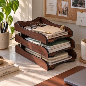 Könnte beinhalten: Ein dreistufiger Schreibtisch-Organizer aus dunklem Holz mit sichtbarer Maserung. Der Organizer verfügt über Metallstützstangen, die jede Ebene verbinden, welche mit verschiedenen Notizbüchern, einer goldenen Büroklammer, einem Stift und einer Brille gefüllt sind.