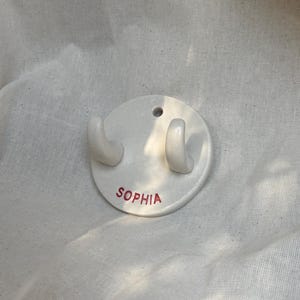 Op de afbeelding: Een ronde witte keramische wandhaak met twee gebogen haken en een klein gat aan de bovenkant voor montage. De naam SOPHIA is in rode hoofdletters gedrukt op het onderste gedeelte van het gladde, glanzende witte oppervlak van de wandhaak.