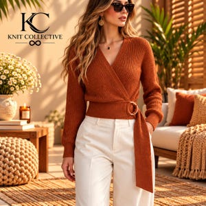 Op de afbeelding: Een roestkleurige, langmouwige, geribbelde gebreide overslagtrui met een zijsluiting met een ronde gesp, gecombineerd met een witte broek. De afbeelding bevat de tekst KC KNIT COLLECTIVE met een oneindigheidssymbool.