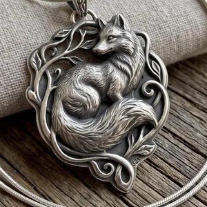 May include: A silver-toned pendant necklace featuring a detailed fox figure curled within a frame of swirling vine-like metalwork. The pendant hangs from a thin, smooth silver-toned chain, showcasing a polished metallic finish with dark recessed areas.