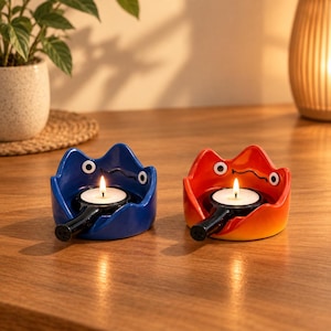 May include: Two ceramic candle holders shaped like stylised creatures with wide eyes and a wavy rim. One holder is deep blue, and the other is a gradient of orange to yellow. Each holds a small black pan containing a lit white tea light candle.