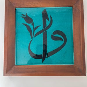 May include: A square ceramic tile featuring a glossy turquoise surface with a black calligraphic design and a tulip motif, encased in a simple brown wooden frame. The artwork displays a stylized Arabic letter combined with a floral element.