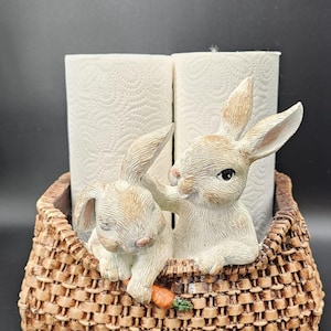 Op de afbeelding: Een decoratieve houder met twee lichtbruine konijnen in een mand met een getextureerde vlechtstructuur. Eén konijn houdt een kleine oranje wortel vast. De mand is ontworpen voor twee rollen keukenpapier die achter de konijnen zichtbaar zijn.