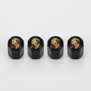 May include: A set of four black tire valve stem caps, each featuring a circular top with the Porsche crest logo in gold, black, and red colors. The caps have a hexagonal base design and a smooth, matte black finish on the cylindrical sides.