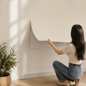 May include: A roll of light beige, textured wallpaper being applied to a wall. The wallpaper has a subtle, woven fabric-like appearance and is partially unrolled, showing a smooth, adhesive backing as it is positioned against the surface.