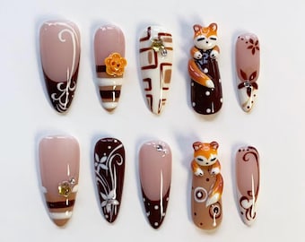May include: A set of ten press-on nails featuring a color palette of nude, dark brown, and orange. Designs include white swirls, horizontal stripes, geometric patterns, floral motifs, small rhinestones, and three-dimensional fox figures.