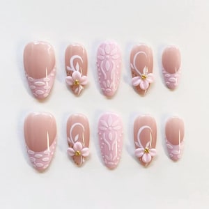 May include: A set of ten press-on nails in varying sizes, featuring a light pink and nude color palette. The designs include raised white floral patterns, white dotted textures, and small three-dimensional pink flowers with gold centers.