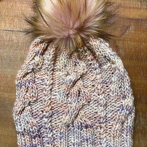 Op de afbeelding: Een gebreide beanie met een kabelbreipatroon in tinten roze, wit en bruin. De beanie heeft een pluche roze en bruine pompon bovenop.
