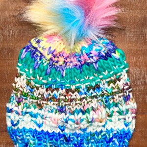 May include: A colourful knitted beanie with a multi-coloured pom pom. The beanie is made with a variety of blue, green, white, and brown yarns.