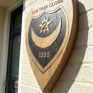 Pode incluir: Uma placa de casa de madeira em forma de escudo com uma borda de madeira natural e um centro escuro. Apresenta o texto ELM TREE CLOSE no topo, o número 2 dentro de uma estrela e o ano 1898 abaixo, esculpidos com um design de lua crescente.