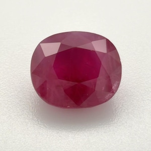 May include: A loose, cushion-cut ruby gemstone with a deep, saturated red colour. The stone features a faceted surface that creates geometric light reflections across its top, showcasing a clear, transparent quality and a polished, smooth finish.