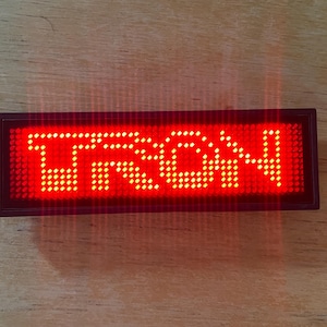 Op de afbeelding: Een rechthoekig LED-display met het woord "TRON" in helderrode, gepixelde letters. Het display is ingekaderd in een donker frame en ligt op een houten oppervlak. De verlichte tekst is het middelpunt.