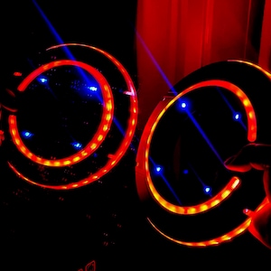 Könnte beinhalten: Zwei beleuchtete, kreisförmige Objekte mit roten und gelben Lichtern sowie blauen Akzentlichtern. Die Objekte werden in Händen vor einem dunklen Hintergrund gehalten, mit roten Lichtstreifen. Das Gesamtthema ist futuristisch oder technologiebezogen.