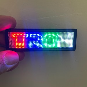 Op de afbeelding: Een rechthoekig LED-display met het woord "TRON" verlicht in rood, blauw, groen en wit. Het display wordt in een hand gehouden. Het apparaat heeft een zwart frame en is waarschijnlijk een naamplaatje of een klein informatiescherm.