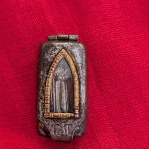 May include: A rectangular metal amulet case with a weathered dark gray finish, featuring a central pointed arch window framed in brass. Inside the window is a relief figure of a standing person. The base of the case displays engraved symbols.
