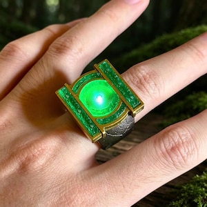 May include: A metallic ring featuring a rectangular gold-toned frame with textured green panels and a central circular element that emits a bright green light. The band of the ring has a dark, weathered, and uneven surface texture.