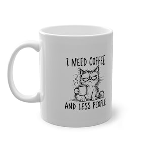 Könnte beinhalten: Ein weißer Keramikbecher mit einer schwarzen Strichzeichnung einer mürrischen Katze, die eine kleine Tasse hält. Der Text über der Katze lautet I NEED COFFEE und der Text unter der Katze lautet AND LESS PEOPLE. Das Motiv ist mittig platziert.