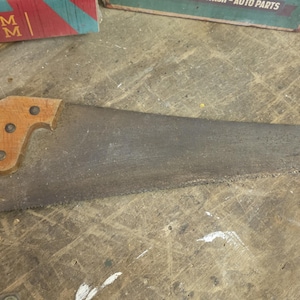 May include: A hand saw featuring a weathered, dark grey metal blade with a serrated edge and a light brown wooden handle secured by three metal rivets. The tool shows signs of age and use with a textured, oxidised surface on the steel blade.