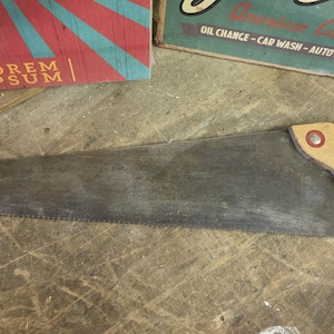 May include: A hand saw with a long, tapered, dark grey metal blade featuring a serrated bottom edge. The handle is made of light-coloured wood and is secured to the blade with three circular fasteners, one of which has a distinct red centre.