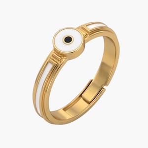 May include: A gold-toned ring featuring a circular focal point with a white enamel background and a central black dot. The band includes white enamel inlay accents along the sides and an adjustable opening at the back for a flexible fit.