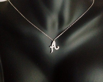 Può includere: Una collana d'argento con un ciondolo a forma di lettera "A". Il ciondolo è tempestato di piccole pietre preziose trasparenti. La collana è esposta su un manichino nero.