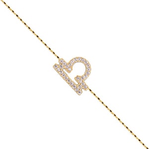 May include: A delicate gold-colored necklace featuring a Libra zodiac symbol. The symbol is encrusted with small, sparkling white gemstones. The chain has a subtle, textured design, and the pendant is positioned diagonally.