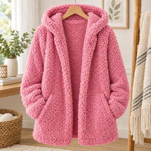 Könnte beinhalten: Eine leuchtend pinke Kapuzenjacke aus einem strukturierten, flauschigen Fleecematerial. Die Jacke verfügt über lange Ärmel, ein offenes Frontdesign und zwei Seitentaschen. Das Innenfutter ist farblich auf das pinke Äußere der Jacke abgestimmt.