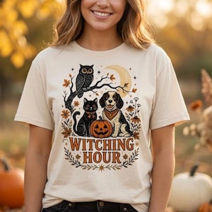 May include: A cream-colored short-sleeve t-shirt featuring a graphic print of an owl on a branch, a black cat, a dog, and a jack-o'-lantern. The design includes autumn leaves, stars, and a crescent moon with the text WITCHING HOUR in orange.