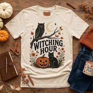 May include: A cream-colored short-sleeve t-shirt featuring a graphic print of an owl on a branch, a black cat, a jack-o'-lantern, and the text "WITCHING HOUR" in a dark font, surrounded by autumn flowers, stars, and a crescent moon.