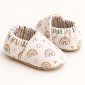Op de afbeelding: Een paar zachte babyschoentjes van gebroken wit textiel met een herhalend patroon van regenbogen in zachte tinten, kleine hartjes en gestippelde cirkels. De schoentjes hebben een elastische opening, een beige zool en een gestreepte voering.