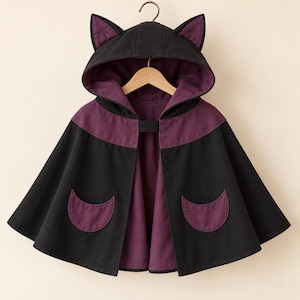 Pode incluir: Uma capa com capuz preta e roxa com um design de orelhas de gato no capuz, forro interior roxo escuro, uma tira de fecho frontal preta e dois bolsos roxos em forma de meia-lua nos painéis frontais pretos da capa.