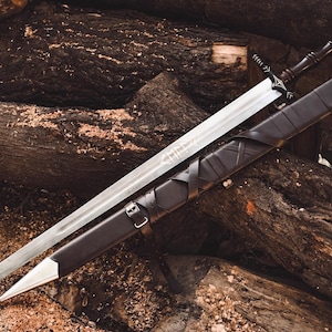 May include: A silver-coloured metal sword with a dark handle featuring white etched detailing. The blade has a central fuller and engraved markings. It is accompanied by a black leather scabbard with a silver metal tip and a buckled strap.