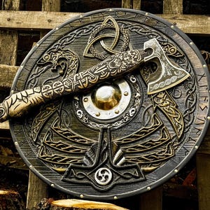 May include: A circular wooden shield featuring intricate carved knotwork, a central metal boss, and runic inscriptions along the rim, paired with a matching hand axe with a detailed carved wooden handle and a metal blade with etched patterns.