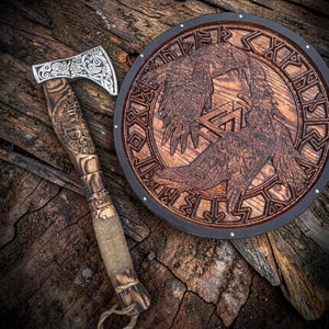 May include: A metal axe with an intricate silver-toned blade and a dark wood handle featuring carved patterns and a rope grip, positioned next to a circular wooden plaque displaying a relief carving of two ravens and runic symbols.