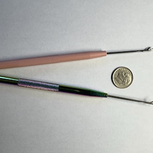 May include: Two crochet hooks with metal tips and textured handles, one pink and one iridescent green. A US dime is next to the hooks for size comparison. The hooks are on a white surface.