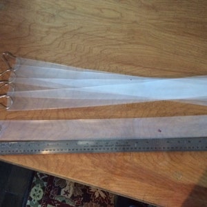 May include: Four clear plastic strips with metal hooks at one end. The strips are laid out on a wooden surface next to a metal ruler.