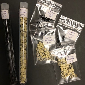 Op de afbeelding: Een assortiment haarverlengingsbenodigdheden. Twee doorzichtige buisjes gevuld met zwarte en blonde micro-kralen, met het label "3mm Black 150" en "3mm Blonde 150". Verschillende kleine plastic zakjes bevatten meer kralen, met het label "3mm Black 50" en "3mm Blonde 50".