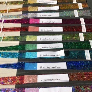 May include: A collection of 13 different colors of shimmering iridescent thread, labeled with their names: sizzling champagne, sizzling gold, sizzling silver, sizzling hot pink, sizzling red, sizzling lime green, sizzling lucky green, sizzling ocean blue, sizzling royal blue, sizzling fuchsia, and sizzling violet.