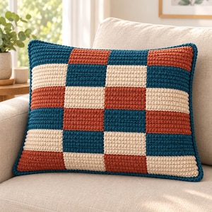 May include: A square crochet pillow cover featuring a repeating checkerboard pattern in deep teal, burnt orange, and cream yarn. The textile construction displays a consistent, textured stitch throughout the entire surface of the cushion.