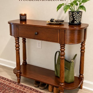 May include: A brown wooden console table with a single drawer, a lower shelf, and turned legs. The text Woodencraftdecorart is visible above the table. A candle, a small tray with keys, and a potted plant sit on the top surface.