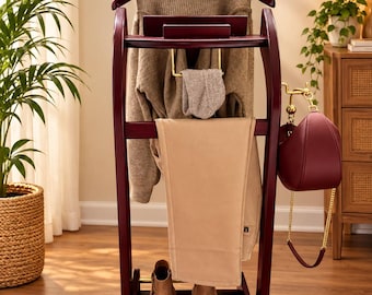 May include: A dark cherry wood valet stand featuring a curved hanger for a beige knit sweater, a horizontal bar for tan trousers, a small tray, a gold-toned metal rack for accessories, and a bottom shelf holding a pair of tan ankle boots.